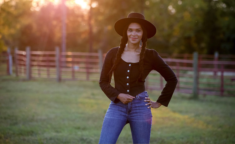 Una fotografía de una mujer vestida con una camisa negra, un sombrero de fieltro negro y unos vaqueros azules, posando en un prado verde.